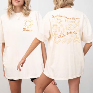 Op de afbeelding: Crèmekleurige t-shirts met goudkleurige afbeeldingen. Eén shirt heeft een zon en het woord "Mama". Het andere shirt heeft de tekst "First trip around the sun" en verjaardagsillustraties, waaronder een raket, taart en ballonnen. De naam "Shai's" is ook zichtbaar.