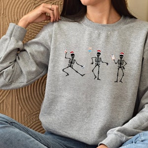 Pode incluir: Um moletom cinza mesclado com um desenho de tr&ecirc;s esqueletos dan&ccedil;ando usando gorros de Papai Noel. Cada esqueleto est&aacute; segurando equipamentos m&eacute;dicos. O desenho &eacute; em preto e vermelho. O moletom tem mangas compridas e gola redonda.
