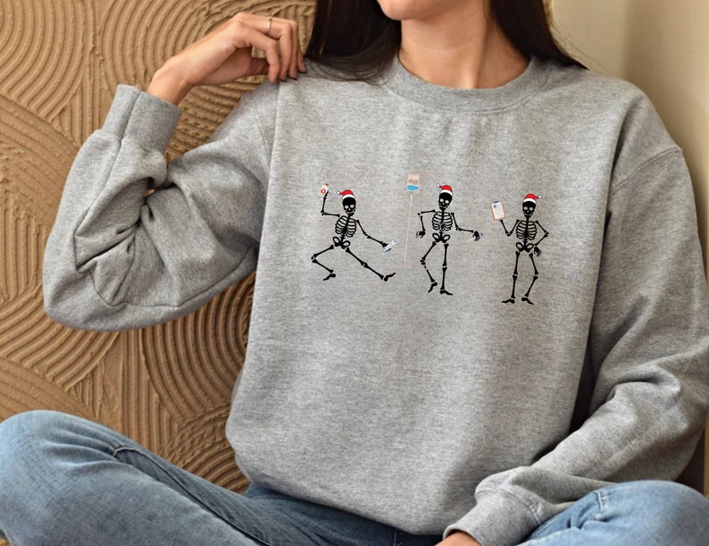 Pode incluir: Um moletom cinza mesclado com um desenho de tr&ecirc;s esqueletos dan&ccedil;ando usando gorros de Papai Noel. Cada esqueleto est&aacute; segurando equipamentos m&eacute;dicos. O desenho &eacute; em preto e vermelho. O moletom tem mangas compridas e gola redonda.