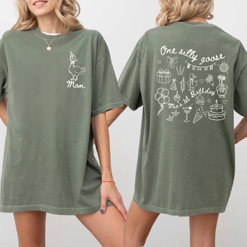 Peut inclure: T-shirt vert olive avec un motif d'oie blanche portant un chapeau de f&ecirc;te et le mot "Mom" sur le devant. Le dos du t-shirt pr&eacute;sente le texte "One silly goose" et des illustrations sur le th&egrave;me de l'anniversaire.