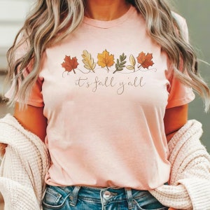 May include: A light pink t-shirt with a fall-themed design. The design features a row of colorful leaves in shades of red, yellow, and orange, with the phrase "it's fall y'all" in a script font.