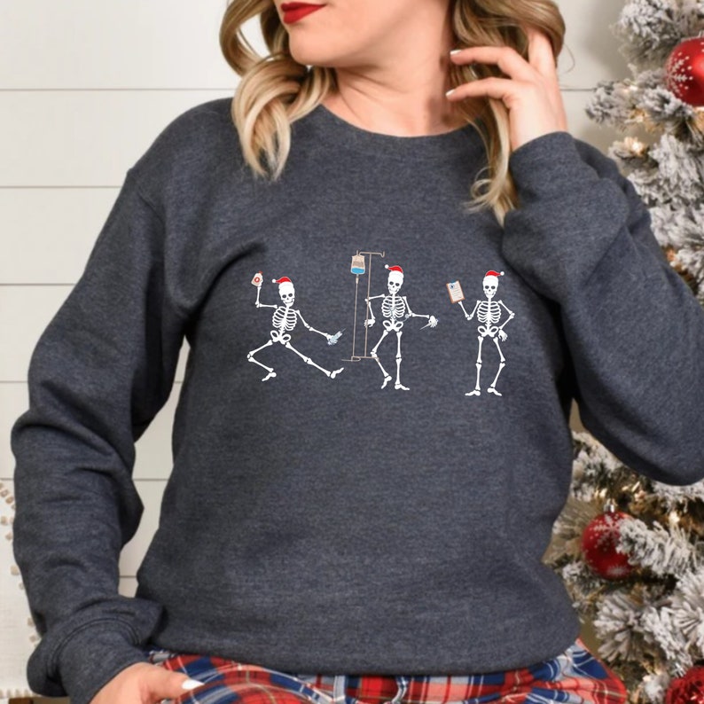 Pode incluir: Camisola cinza escuro com um gr&aacute;fico de tr&ecirc;s figuras de esqueleto usando chap&eacute;us de Papai Noel. Um esqueleto est&aacute; segurando uma seringa, outro um gotejamento intravenoso e o terceiro um prontu&aacute;rio m&eacute;dico. Os esqueletos est&atilde;o em uma pose de dan&ccedil;a.