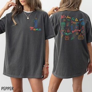 May include: Dark gray t-shirt with a colorful design. The front features a number one candle and the word "Mama". The back of the shirt has the text "It's fun to be one" and a birthday-themed graphic. The shirt is a relaxed fit.