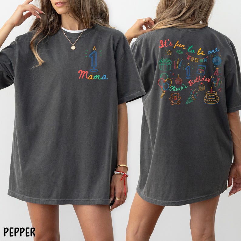 May include: Dark gray t-shirt with a colorful design. The front features a number one candle and the word "Mama". The back of the shirt has the text "It's fun to be one" and a birthday-themed graphic. The shirt is a relaxed fit.