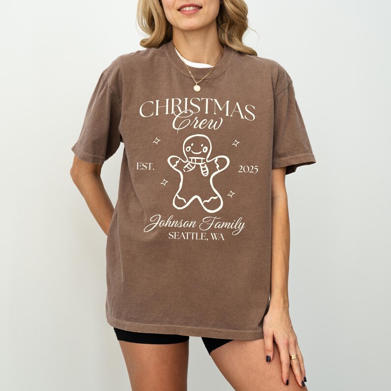 May include: A brown t-shirt with the text "CHRISTMAS CREW" and a gingerbread man graphic. Below the graphic is "Johnson Family, Seattle, WA" and the year "2025". The shirt has a crew neck and short sleeves.
