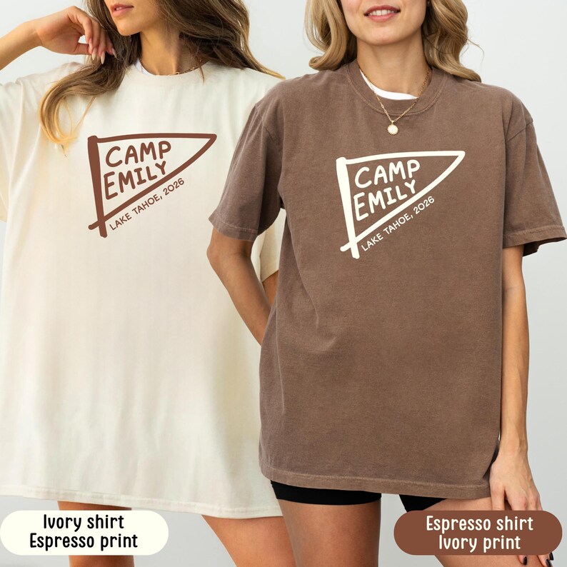 May include: Two short-sleeve crewneck t-shirts. One is ivory with an espresso brown graphic of a pennant flag featuring the text CAMP EMILY LAKE TAHOE, 2026. The other is espresso brown with an ivory graphic of the same pennant flag and text.