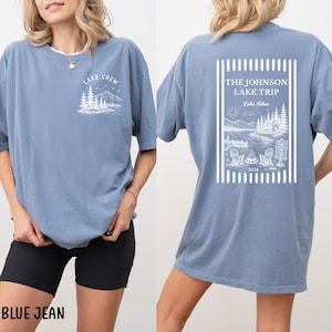 Puede incluir: Camiseta azul vaquero con dise&ntilde;os gr&aacute;ficos blancos. La parte delantera presenta "LAKE CREW" sobre una escena de monta&ntilde;a y &aacute;rboles. La parte trasera muestra "THE JOHNSON LAKE TRIP" con una escena de lago, sillas y el a&ntilde;o 2026.
