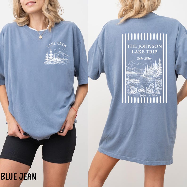 Puede incluir: Camiseta azul vaquero con dise&ntilde;os gr&aacute;ficos blancos. La parte delantera presenta "LAKE CREW" sobre una escena de monta&ntilde;a y &aacute;rboles. La parte trasera muestra "THE JOHNSON LAKE TRIP" con una escena de lago, sillas y el a&ntilde;o 2026.