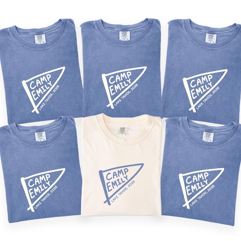 May include: Five blue t-shirts and one cream t-shirt, each featuring a white pennant graphic with the text "CAMP EMILY" and "LAKE TAHOE, 2026". The cream shirt features the same pennant graphic in blue. All shirts have a crew neck design.