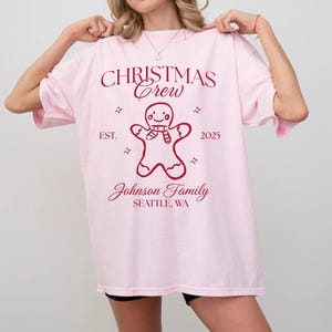 May include: A light pink t-shirt with the text "CHRISTMAS Crew" and "EST. 2025" in burgundy. A gingerbread man graphic is centered below the text. The words "Johnson Family" and "SEATTLE, WA" are printed below the gingerbread man.