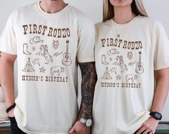 May include: Two cream-colored t-shirts with brown text and illustrations. The text on the shirts reads "First Rodeo" and "Hudson's Birthday". The illustrations include a cowboy hat, boots, a horse, a guitar, a tent, and fireworks.