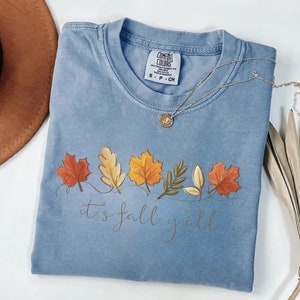 May include: A light blue Comfort Colors t-shirt with a fall leaves design and the text "it's fall y'all". The shirt has a crew neck and short sleeves. A gold necklace with a pendant is draped across the shirt.