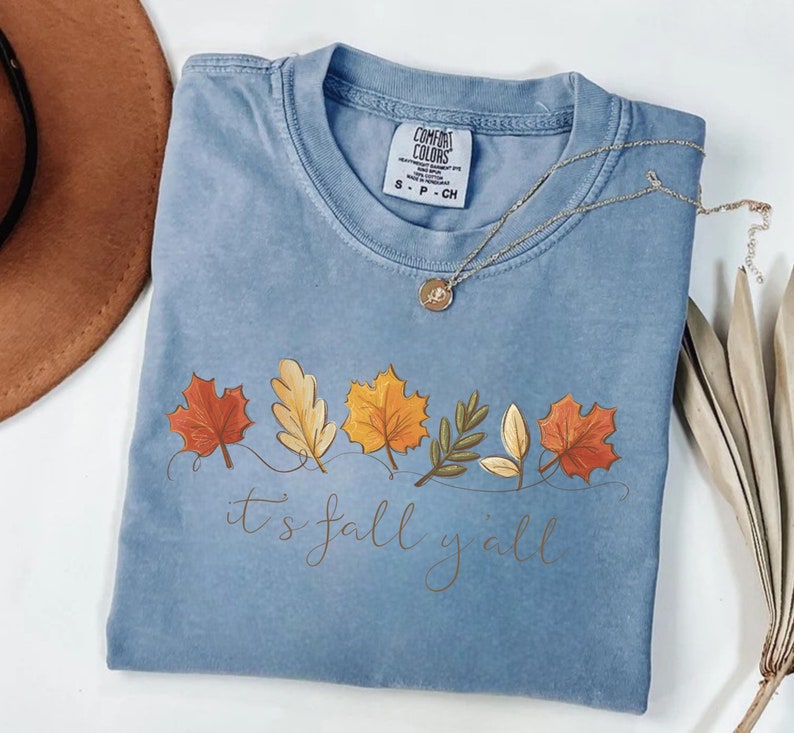 May include: A light blue Comfort Colors t-shirt with a fall leaves design and the text "it's fall y'all". The shirt has a crew neck and short sleeves. A gold necklace with a pendant is draped across the shirt.