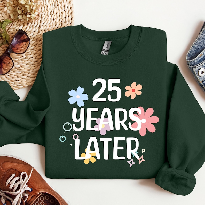 May include: A dark green sweatshirt with white text that reads "25 years later" surrounded by colourful flowers.