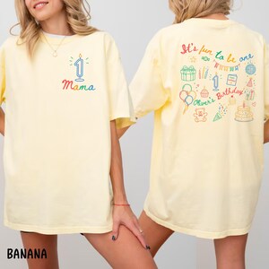 May include: A light yellow t-shirt with the word "Mama" and a number one candle graphic on the front. The back of the shirt has the text "It's fun to be one" with birthday-themed illustrations. The shirt is a casual style.