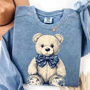 Könnte beinhalten: Ein blaues Sweatshirt mit einem Comic-Teddybär, der eine blaue Schleife mit Blumenmuster trägt. Der Bär sitzt mit seinen Pfoten vor sich.