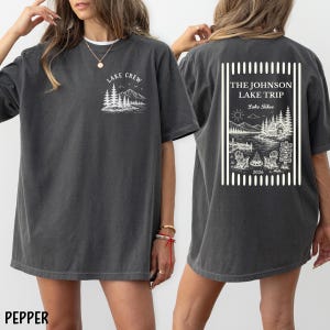 Puede incluir: Camiseta color pimienta con un gr&aacute;fico blanco en la parte delantera que dice "Lake Crew" con un dise&ntilde;o de monta&ntilde;a y &aacute;rboles. La parte posterior de la camisa tiene un dise&ntilde;o rectangular con el texto "The Johnson Lake Trip Lake Tahoe 2026".