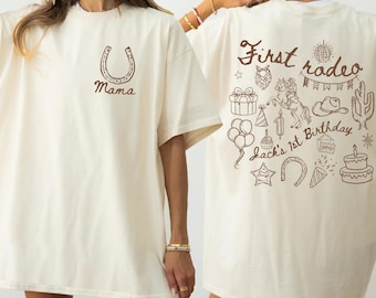 May include: Ivory-colored t-shirt with a horseshoe and the word "Mama" printed in brown. The back of the shirt features a "First rodeo" design with a horse, cake, balloons, and other rodeo-themed illustrations. "Jack's 1st Birthday" is also printed on the back.