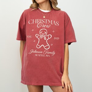 May include: A red t-shirt with the text "CHRISTMAS Crew" and a gingerbread man graphic. The shirt also includes the text "EST. 2025" and "Johnson Family SEATTLE, WA". The shirt is a solid red color.