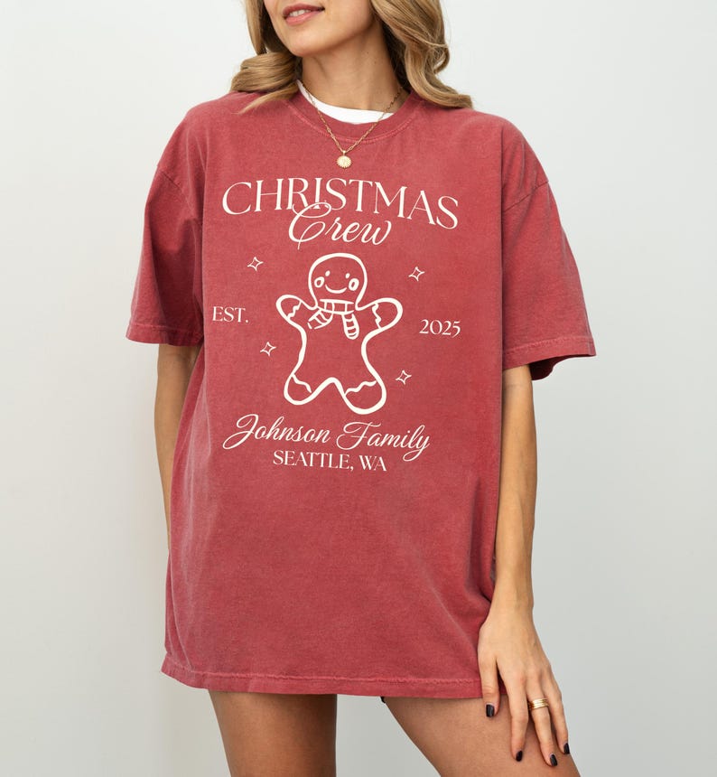 May include: A red t-shirt with the text "CHRISTMAS Crew" and a gingerbread man graphic. The shirt also includes the text "EST. 2025" and "Johnson Family SEATTLE, WA". The shirt is a solid red color.