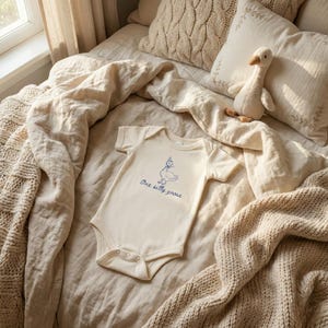 Peut inclure: Body b&eacute;b&eacute; cr&egrave;me avec une illustration bleue d'une oie portant un chapeau et le texte "One silly goose". Le body est pos&eacute; sur un lit avec une couverture en tricot, des oreillers et un jouet en forme d'oie.