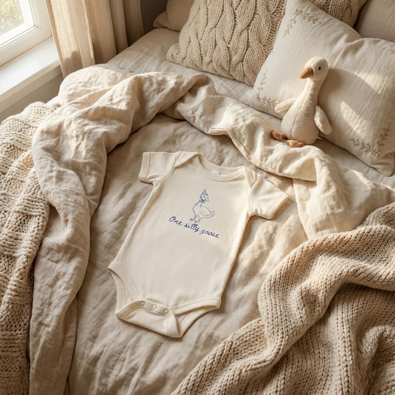 Peut inclure: Body b&eacute;b&eacute; cr&egrave;me avec une illustration bleue d'une oie portant un chapeau et le texte "One silly goose". Le body est pos&eacute; sur un lit avec une couverture en tricot, des oreillers et un jouet en forme d'oie.
