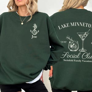 May include: Dark green sweatshirt with white graphic designs. The front features an anchor and the name "Joe". The back displays "Lake Minnetonka Social Club" with cocktail illustrations, "August 2025", and "Steinfield Family Vacation".