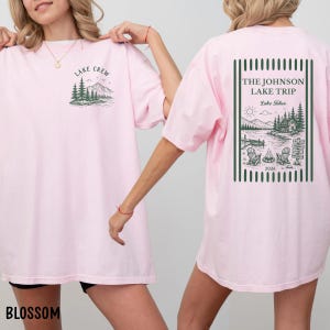 Puede incluir: Camisetas de color rosa claro con gr&aacute;ficos delanteros y traseros. El gr&aacute;fico frontal dice "Lake Crew" con un dise&ntilde;o de monta&ntilde;a y &aacute;rboles. El gr&aacute;fico trasero dice "The Johnson Lake Trip" con una escena de lago, sillas y el a&ntilde;o 2026.