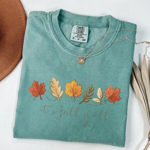 May include: A teal Comfort Colors t-shirt with a fall leaves design and the text "it's fall y'all". The design features colorful leaves in shades of red, orange, and yellow. A silver necklace with a pendant is on the shirt.