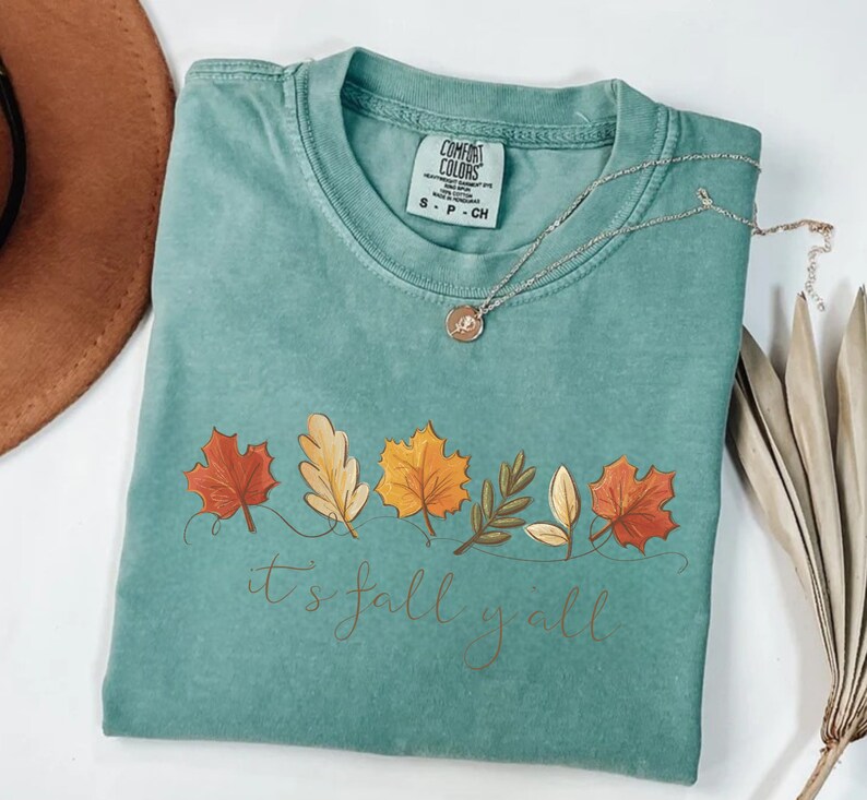 May include: A teal Comfort Colors t-shirt with a fall leaves design and the text "it's fall y'all". The design features colorful leaves in shades of red, orange, and yellow. A silver necklace with a pendant is on the shirt.