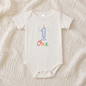 May include: A cream-colored baby onesie with a blue number one candle graphic. The candle has a flame and is surrounded by green and yellow accents. The word "One" is written in colorful script below the candle.