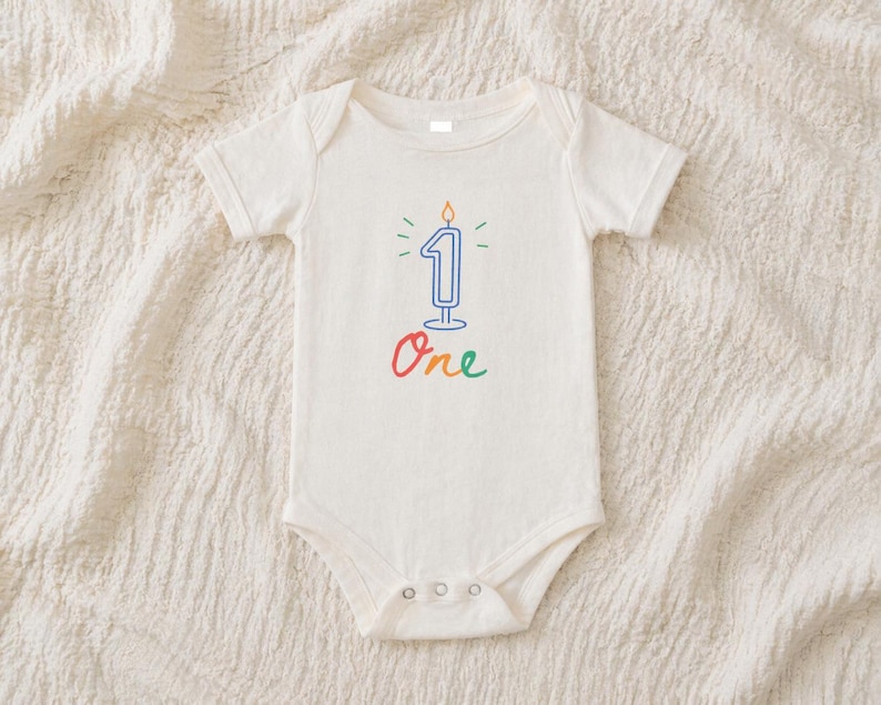 May include: A cream-colored baby onesie with a blue number one candle graphic. The candle has a flame and is surrounded by green and yellow accents. The word "One" is written in colorful script below the candle.
