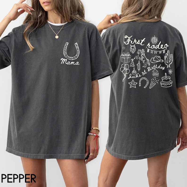 K&ouml;nnte beinhalten: Pfefferfarbenes T-Shirt mit einem wei&szlig;en Hufeisen und dem Wort "Mama" auf der Vorderseite. Auf der R&uuml;ckseite des Shirts steht der Text "First rodeo" und verschiedene Rodeo-Illustrationen, darunter ein Kuchen und ein Kaktus.