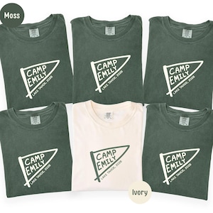 May include: Five moss green t-shirts and one ivory t-shirt, each featuring a white pennant graphic with the text CAMP EMILY and LAKE TAHOE, 2026. The shirts have a crew neckline and a visible brand label on the inside of the collar area.