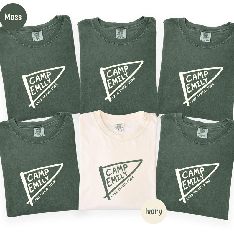 May include: Five moss green t-shirts and one ivory t-shirt, each featuring a white pennant graphic with the text CAMP EMILY and LAKE TAHOE, 2026. The shirts have a crew neckline and a visible brand label on the inside of the collar area.
