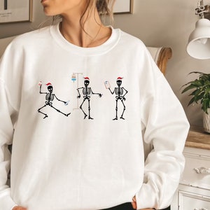 Pode incluir: Camisola branca com um desenho de tr&ecirc;s esqueletos a dan&ccedil;ar com gorros de Pai Natal. Os esqueletos s&atilde;o representados com equipamento m&eacute;dico, incluindo um gotejamento intravenoso e uma prancheta. O desenho &eacute; em preto e vermelho.