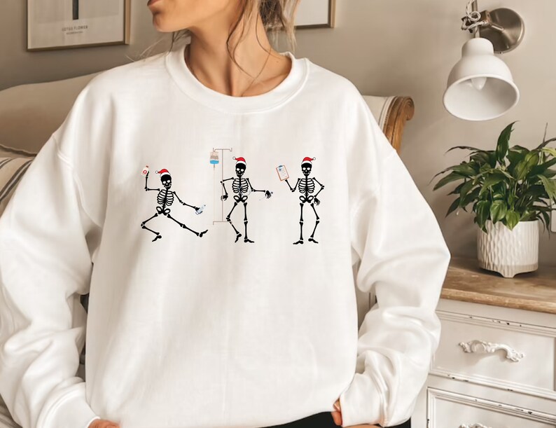 Pode incluir: Camisola branca com um desenho de tr&ecirc;s esqueletos a dan&ccedil;ar com gorros de Pai Natal. Os esqueletos s&atilde;o representados com equipamento m&eacute;dico, incluindo um gotejamento intravenoso e uma prancheta. O desenho &eacute; em preto e vermelho.