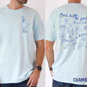 Peut inclure: T-shirt bleu clair avec un dessin d'oie bleue portant un chapeau de f&ecirc;te et le mot "Dad". Le dos du t-shirt porte l'inscription "One silly goose Orlando's 1st Birthday" avec des illustrations sur le th&egrave;me de l'anniversaire. Le mot "CHAMBRAY" est en bas.