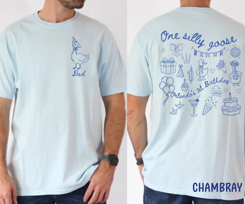Peut inclure: T-shirt bleu clair avec un dessin d'oie bleue portant un chapeau de f&ecirc;te et le mot "Dad". Le dos du t-shirt porte l'inscription "One silly goose Orlando's 1st Birthday" avec des illustrations sur le th&egrave;me de l'anniversaire. Le mot "CHAMBRAY" est en bas.