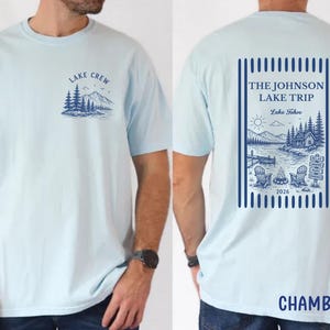 Puede incluir: Camiseta azul claro con un gr&aacute;fico de la orilla del lago en la parte delantera y trasera. La parte delantera presenta un dise&ntilde;o de monta&ntilde;a y &aacute;rboles con el texto "LAKE CREW". La parte trasera muestra una escena del lago con el texto "THE JOHNSON LAKE TRIP Lake Tahoe 2026".