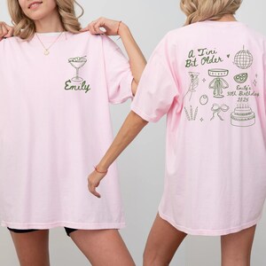 May include: Pink t-shirts with green graphics. One shirt has a martini glass and the name "Emily". The other shirt has the text "A Tiny Bit Older" and birthday-themed illustrations, including a cake and disco ball. The shirts are casual wear.