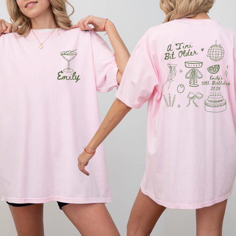 May include: Pink t-shirts with green graphics. One shirt has a martini glass and the name "Emily". The other shirt has the text "A Tiny Bit Older" and birthday-themed illustrations, including a cake and disco ball. The shirts are casual wear.