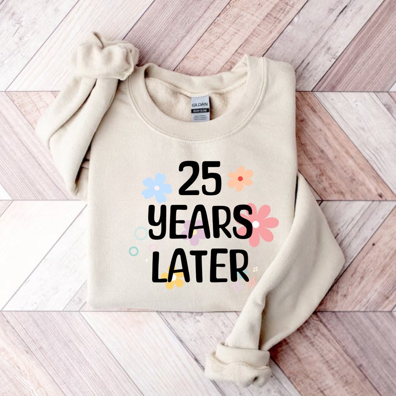 May include: A beige sweatshirt with the text "25 years later" in black lettering and colourful flowers surrounding the text.