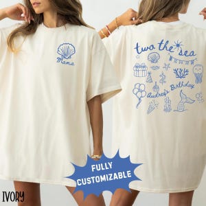 Könnte beinhalten: Elfenbeinfarbenes T-Shirt mit blauer Muschel und "Mama"-Text auf der Vorderseite. Die Rückseite zeigt den Text "two the sea" und Illustrationen zum Thema Meer, darunter eine Meerjungfrau, Luftballons und eine Geburtstagstorte. Vollständig anpassbar.