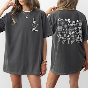 Peut inclure: T-shirt gris fonc&eacute; avec une oie blanche portant un chapeau de f&ecirc;te et le mot "Mom" sur le devant. Le dos du t-shirt pr&eacute;sente le texte "One silly goose" et des illustrations sur le th&egrave;me de l'anniversaire, dont un g&acirc;teau et des ballons.