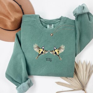 May include: A green sweatshirt with a graphic of two birds facing each other with the word "tits" printed below.