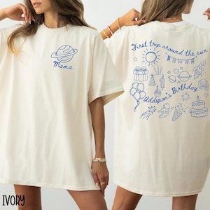 Op de afbeelding: Ivoor T-shirt met een blauwe planeet en "Mama"-afbeelding op de voorkant. De achterkant heeft een blauwe illustratie met de tekst "First trip around the sun" en "Addyson's Birthday" met verjaardagsgerelateerde tekeningen.