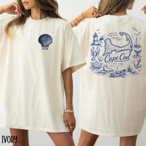 May include: An ivory t-shirt featuring a blue graphic of a seashell and the text 2026 on the front. The back displays a detailed blue illustration of Cape Cod, a lighthouse, a lobster, a seal, a boat, and the text Cape Cod July 2026 Massachusetts.