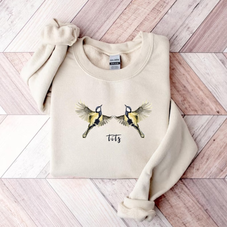 May include: A beige sweatshirt with a graphic of two birds flying with the word "tits" printed below them.
