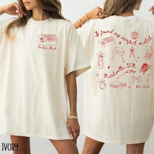 Könnte beinhalten: Elfenbeinfarbenes T-Shirt mit rotem Text und Grafiken. Auf der Vorderseite befindet sich ein Streichholzschachtel-Design mit "PERFECT MATCH" und "Emily's Bach." Auf der Rückseite steht "I found my perfect match" mit verschiedenen Illustrationen, darunter eine Discokugel, Weingläser und ein Ring. Der Text "Nashville, IN" und "Est. 2026" sind ebenfalls vorhanden.